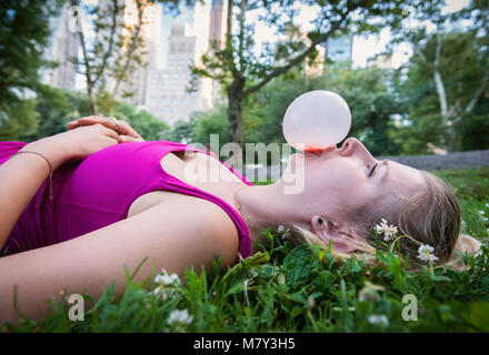 Una donna che stabilisce in erba e soffiando una bolla con la gomma in Central Park di New York City Foto Stock