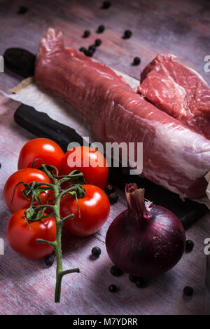 Foto verticale di tutto il filetto di maiale. La carne cruda è su vintage tavola di legno con Red Cherry Pomodoro e cipolla viola. Bacche di ginepro sono fuoriuscite di ar Foto Stock