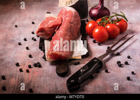 Foto orizzontale di tutto il filetto di maiale. La carne cruda è su vintage tagliere in legno con grande forcella e pochi rossi pomodori ciliegini. Bacche di ginepro sono versate Foto Stock