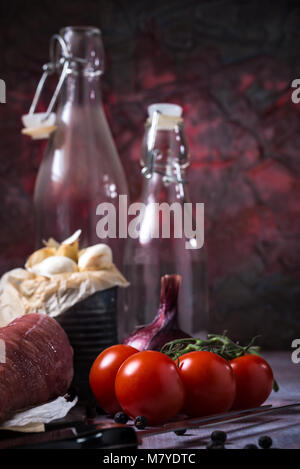 Foto verticale di alcuni rossi pomodori ciliegini. Il vegetale è collocato su vintage tavola di legno accanto alle materie di filetti di maiale fresche, viola la cipolla e il vecchio può Foto Stock