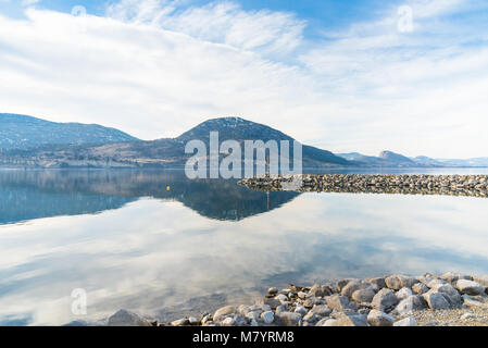 Lago calmo che riflette il cielo blu e nuvole con frangiflutti e le montagne in distanza Foto Stock