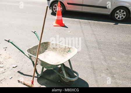 Immagine di rustica costruzione sporco carriola con push ginestra sulla strada, arancione per la costruzione di strade di cono in background. Foto Stock