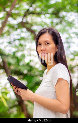 Ritratto di giovane sorridente donna asiatica con un cellulare in un parco verde che guarda verso la telecamera Foto Stock