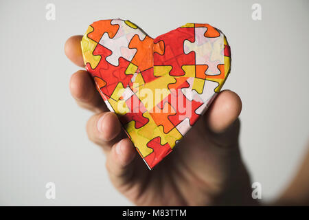 Clousep di un giovane uomo caucasico tenendo un cuore modellato con molti pezzi di un puzzle di diversi colori, simbolo della sensibilizzazione sull'autismo Foto Stock