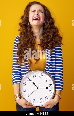 Triste donna elegante con lunga ondulato brunette capelli isolati su sfondo giallo con orologio Foto Stock