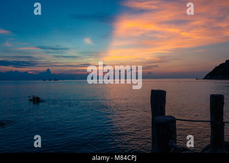 Incredibile tramonto sul mare con metà metà blu rosa e giallo cielo, dei bastoni di legno in primo piano e due persone sagome su un kayak Foto Stock