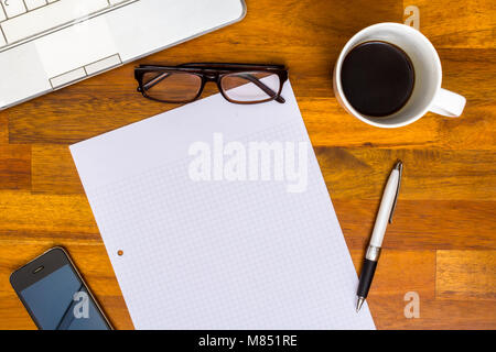 In legno marrone orking scrivania con computer portatile, collegio nota blocco carta, penne, telefono cellulare, bicchieri, Caffè Nero Foto Stock