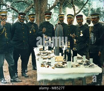 Foto colorata del conte generale giapponese Nogi Maresuke (1849 - 1912) (centro) e ufficiali che si levano in piedi intorno ad un tavolo durante una celebrazione della vittoria durante la guerra russo-giapponese, Port Arthur, Cina, 1905. La proposta è riempita con bottiglie di piatti di cibo, così come un guscio di Malta. (Foto di Burton Holmes) Foto Stock