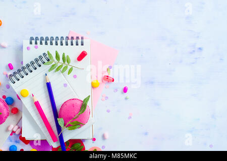 Rivestito in bianco quaderni con matite colorate, dolci e rosa macaron cookie su uno sfondo luminoso con copia spazio. Pianificazione del partito concetto di divertimento. Foto Stock