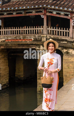 Una bella ragazza in un bianco tradizionale abito lungo sorge accanto a Chùa Cầu ponte giapponese, un edificio del XVIII secolo in legno intagliato ponte che incorpora un santuario, a Hoi An, Vietnam Foto Stock