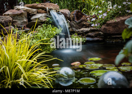 Circondato da fiori e vegetazione lussureggiante, una cascata di seta fluisce fuori delle rocce in un grazioso stagno pieno di pesci rossi Foto Stock