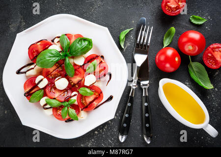 Insalata caprese. Pomodori, mozzarella, pomodoro, olio di oliva, basilico foglie di erbe e salsa balsamica su sfondo scuro. Il cibo italiano. Vista superiore Foto Stock