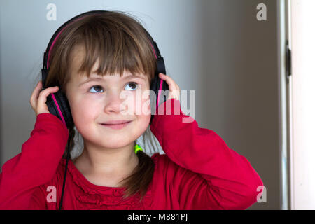 Foto di felice bambina carina bambina con grandi cuffie a casa. Gioiosa ragazza bambino ascoltare musica su grigio sfondo sfocato. Home, technolo Foto Stock