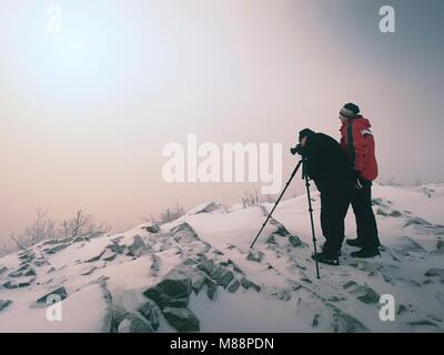 Gli escursionisti e appassionati di fotografia soggiorno sul picco innevato al treppiede. Gli uomini sulla scogliera di parlare e pensare. Inverno da sogno paesaggio con misty sunrise Foto Stock