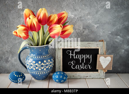 Rosso-tulipani gialli in ceramica blu brocca con uova di Pasqua e di una lavagna nera su sfondo grigio. Testo 'Felice Pasqua" sulla scheda. Foto Stock