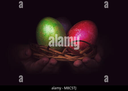 Close up di colorate uova di Pasqua in un cesto di uova dipinte nel bambino la mano di notte su sfondo nero. in verde e rosso Foto Stock
