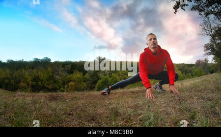 Un giovane forte athletic Uomo in camicia rossa con un cappuccio e nero sport leggins esegue la fase di riscaldamento con le gambe prima di fare jogging su una foresta verde backgro Foto Stock