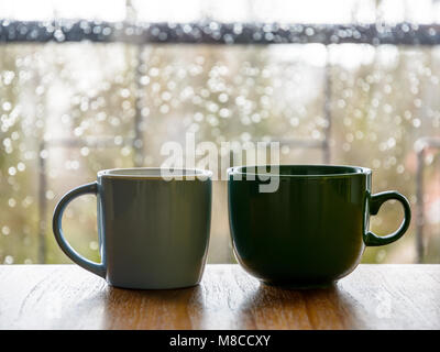 Due tazze su una tavola di legno di fronte a una finestra di vetro ricoperta con gocce di pioggia. Foto Stock