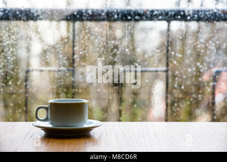 Una tazza da caffè bianca su un tavolo di legno davanti a una finestra di vetro coperta da gocce di pioggia con una profondità di campo poco profonda. Foto Stock
