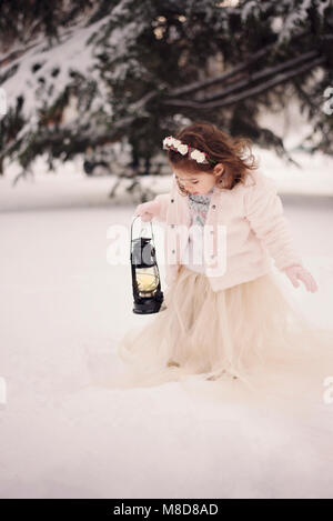 Bambina indossa un cappotto beige e abiti lunghi, guardando la lampada con candela in piedi tra i rami, coperte di neve nel Parco in inverno Foto Stock