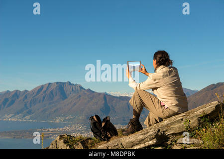 Donna di scattare le foto con la sua tavoletta digitale da una montagna in Ticino, Svizzera Foto Stock