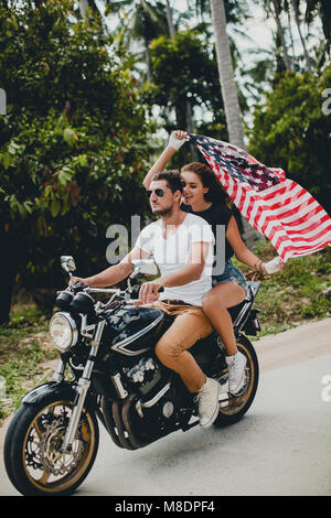 Coppia giovane mantenendo la bandiera americana mentre cavalcate motocicletta sulla strada rurale, Krabi, Thailandia Foto Stock