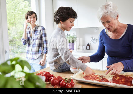 Nonna e nipote rendendo la pizza in cucina, madre in background utilizza lo smartphone Foto Stock