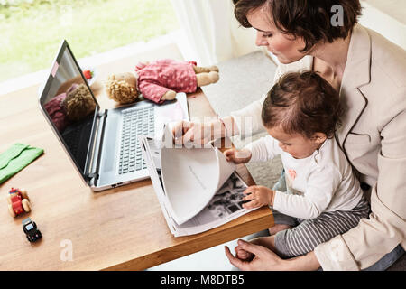 Madre seduta con la nostra bambina, guardando attraverso la documentazione sul computer portatile sul tavolo di fronte a lei Foto Stock