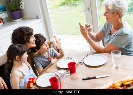 Nonna seduta in cucina, fotografando cresciuta figlia e nipoti, utilizza lo smartphone Foto Stock