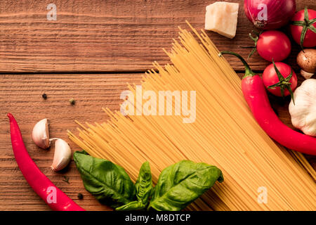 Italian pasta secca spaghetti linguini sparse con pomodori freschi di ciliegia, pepe rosso piccante e il verde del basilico foglie su sfondo di legno. Vista dall'alto. Co Foto Stock