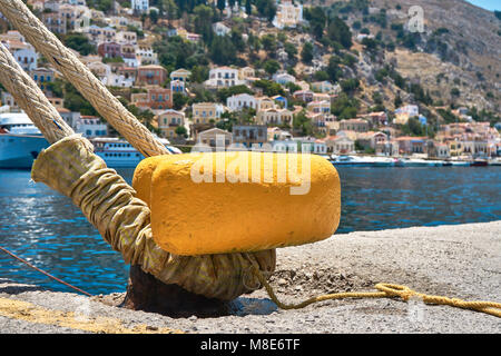 Bitt ormeggio giallo con corda su molo di cemento grigio in acque blu di mare con piccole onde contro case multicolore dell'Isola di Symi Foto Stock