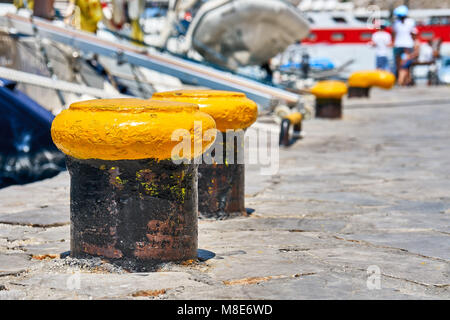 Arrugginito giallo metallo ormeggi situato lungo il molo di cemento grigio contro vascelli sfocati sotto luce solare brillante estate primo piano Foto Stock