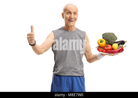 Allegro uomo anziano in azienda di abbigliamento sportivo di una piastra con la frutta e la verdura e la realizzazione di un pollice in alto gesto isolato su sfondo bianco Foto Stock