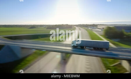 Riprese aeree del carrello bianco con semi rimorchio spostando su autostrada. Sullo sfondo i magazzini e Area Rurale, Sun è l'impostazione. Motion Blur. Foto Stock