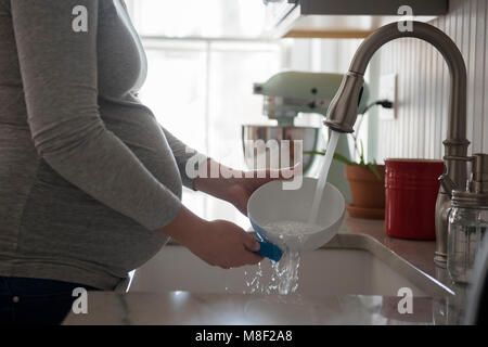 Donna incinta recipiente di lavaggio nel lavello da cucina, metà sezione Foto Stock