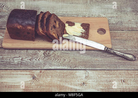 Dark pane di segale con albicocche secche e uva passa. Pane sano. Fette, coltello da tavola e burro. Sfondo di legno Foto Stock