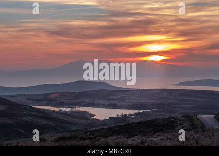 Un tramonto firey causando insolito riflessioni sulla brughiera come il sole tramonta dietro una nebbiosa Arran. L'immagine ha un cupo etherial look. Foto Stock