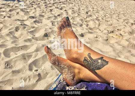 Donna di sabbia piedi incrociati con concia tatuaggio in una sedia a sdraio in spiaggia Foto Stock