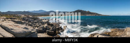 Vista panoramica di lavaggio onde su rocce vicino alla spiaggia di Algajola nella regione della Balagne in Corsica sotto un profondo cielo blu con montagne innevate in th Foto Stock