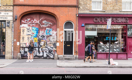 La gente a piedi nella parte anteriore di un vecchio sportello coperto in adesivi e arte di strada murales accanto alla prole store nella strada commerciale, Spitalfields, Shoreditc Foto Stock