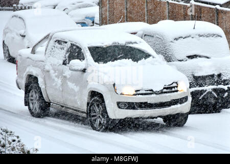 Bianco auto coperto di neve con fari anteriori sul passato di guida dei veicoli parcheggiati in inverno la neve cade sulla coperta di neve road Brentwood Essex England Regno Unito Foto Stock