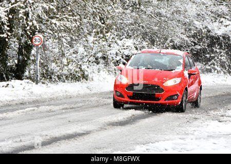 Red Ford auto guidando lungo 30mph coperta di neve country road durante nevicate invernali la creazione di alberi innevati sullo sfondo della scena Inghilterra Essex REGNO UNITO Foto Stock