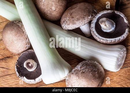 Il verde dei Porri e big marrone di funghi champignon sul tagliere di legno da sopra lo sfondo vegetale Foto Stock