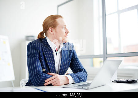 Imprenditore serio seduto al suo posto di lavoro e pensare a qualcosa di Foto Stock