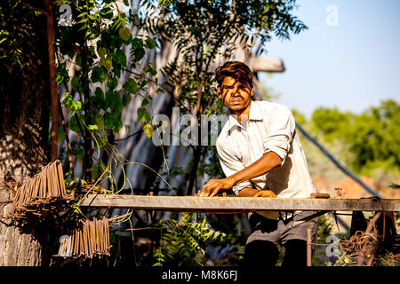 Un bel giovane uomo sta lavorando all' aperto nel villaggio indiano Nipal, piegatura di tondini di ferro durante la bella giornata di sole sotto gli alberi. La luce è golden caldo come Foto Stock