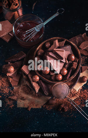 Ciotola di legno con cioccolatini fatti in casa e pezzetti di cioccolato, vetri con una frusta, sparse la polvere di cacao su uno sfondo scuro. Ingredienti Dessert clos Foto Stock