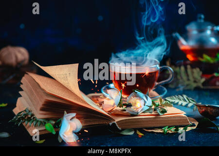 Fumante tazza di tè su un libro aperto con la masterizzazione di magici fiori. Fantasia il concetto di lettura. Dark ancora in vita con copia spazio. Foto Stock