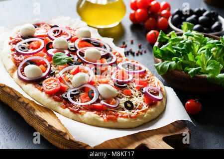 La pizza fatta in casa prima di forno. Pizza con salsa di pomodoro, pomodori, mozzarella, olive nere e cipolla su legno pizza board, primo piano Foto Stock