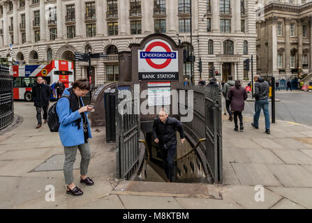 Una donna controllando il suo telefono al di fuori della Banca stazione di tubo di ingresso nella città di Londra come un uomo si precipita il passi dalla stazione della metropolitana di ingresso Foto Stock