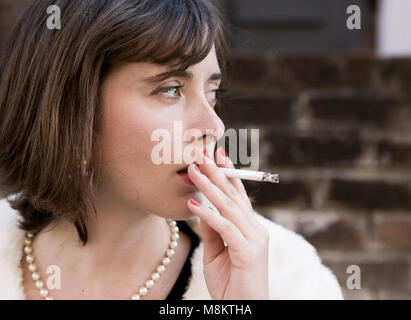 Un ben vestito donna che indossa un rossetto rosso e una collana di perle con una sigaretta accesa nella sua labbra. Foto Stock